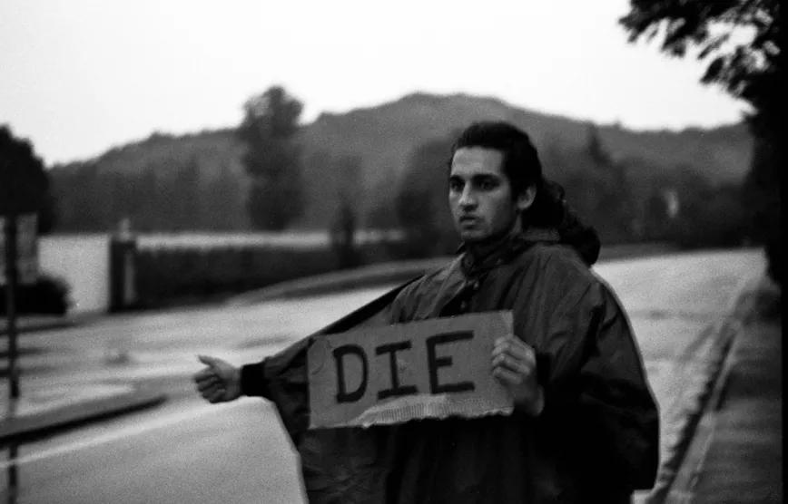 Joven haciendo autoestop con un cartel en el que aparece escrito "Die"
